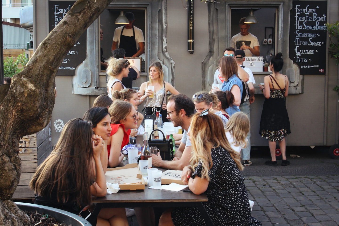 Junge Erwachsene genießen Essen und Gespräche an Picknicktischen in der Nähe von Food Trucks.