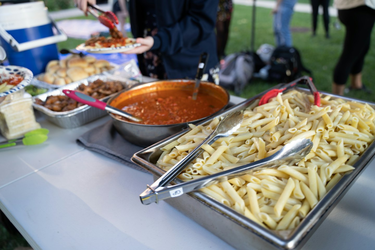 Person reicht Nudeln an einem Buffet-Tisch im Freien.